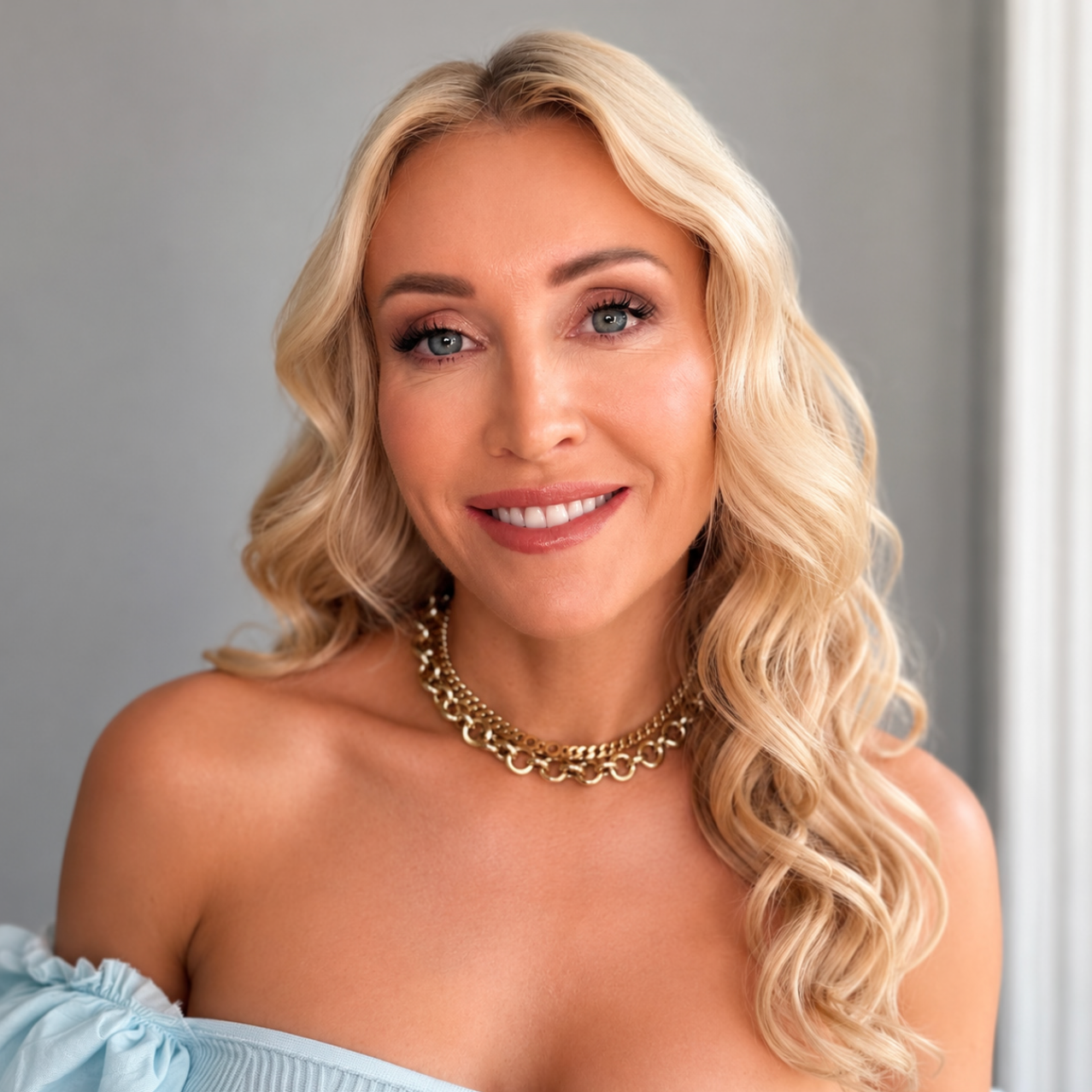 Woman with blonde hair and a gold necklace against a gray background