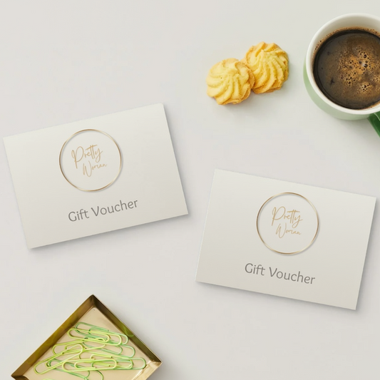 Two white gift voucher cards with gold foil 'Pretty Woman' logos and 'Gift Voucher' text, arranged on a light gray surface with a cup of coffee, cookies, and paperclips nearby.